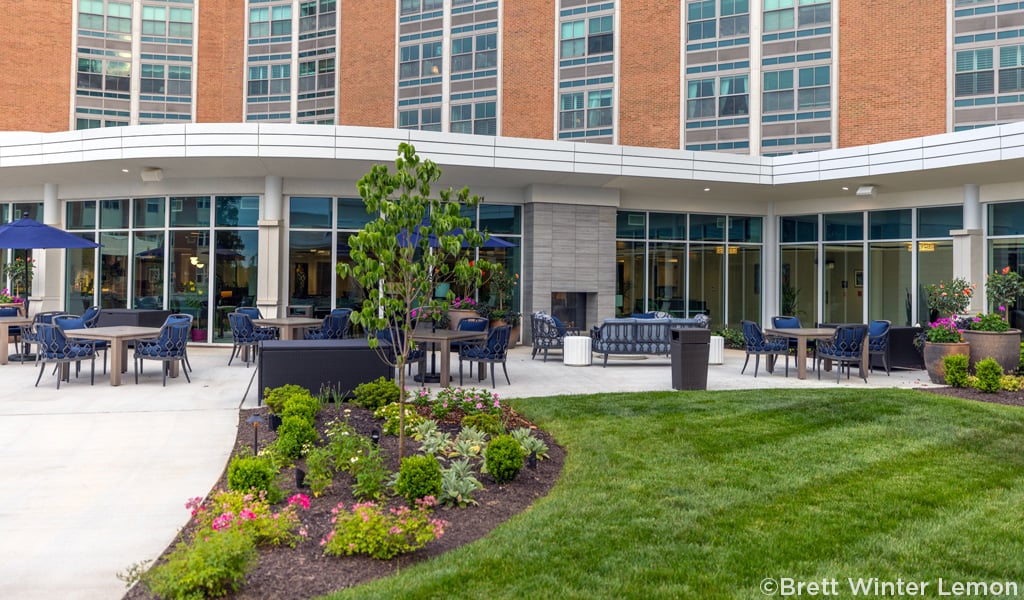 Courtyard seating area with tables, chairs, and a garden in the foreground of a brick senior living facility 