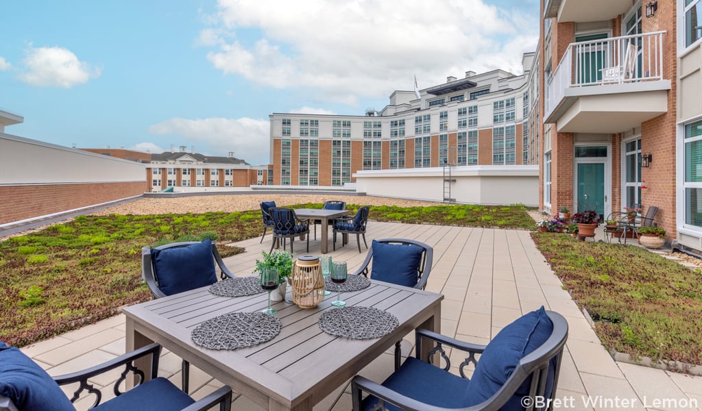 Terrace with 2 tables and blue chairs in an open space adjacent to a brick senior living facility