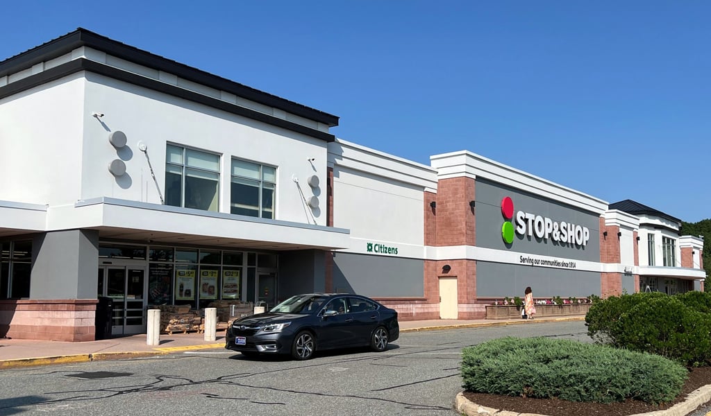 Front entry of a Stop and Shop on a summer day with a blue sky, shopper walking,, and black car driving by the entry. 