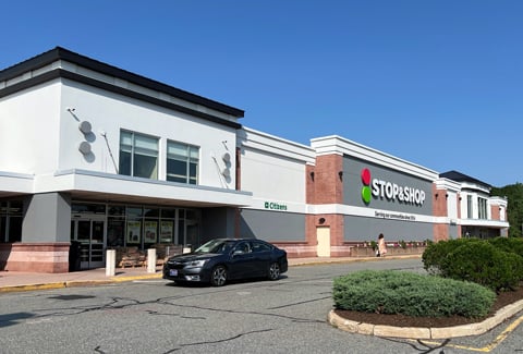 Front entry of a Stop and Shop on a summer day with a blue sky, shopper walking,, and black car driving by the entry. 