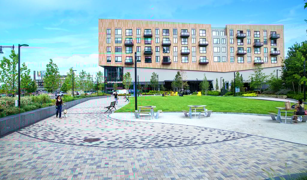 A park in the foreground of a 4-story residential building being actively used by dog walkers and local residents. 