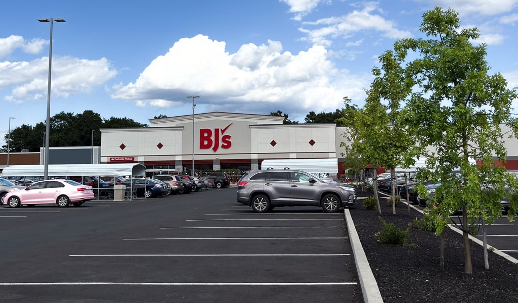 BJ's Wholesale Club storefront with parking lot, landscaped trees, and parked cars under a bright blue sky with clouds.