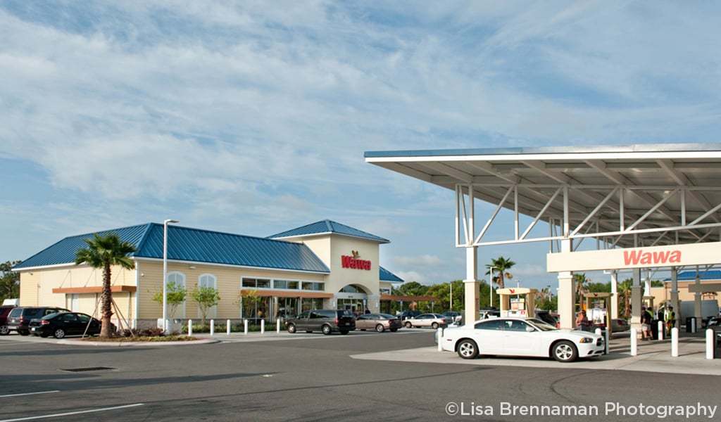 Wawa store with a blue roof, gas pumps, and several parked cars under a partly cloudy sky.