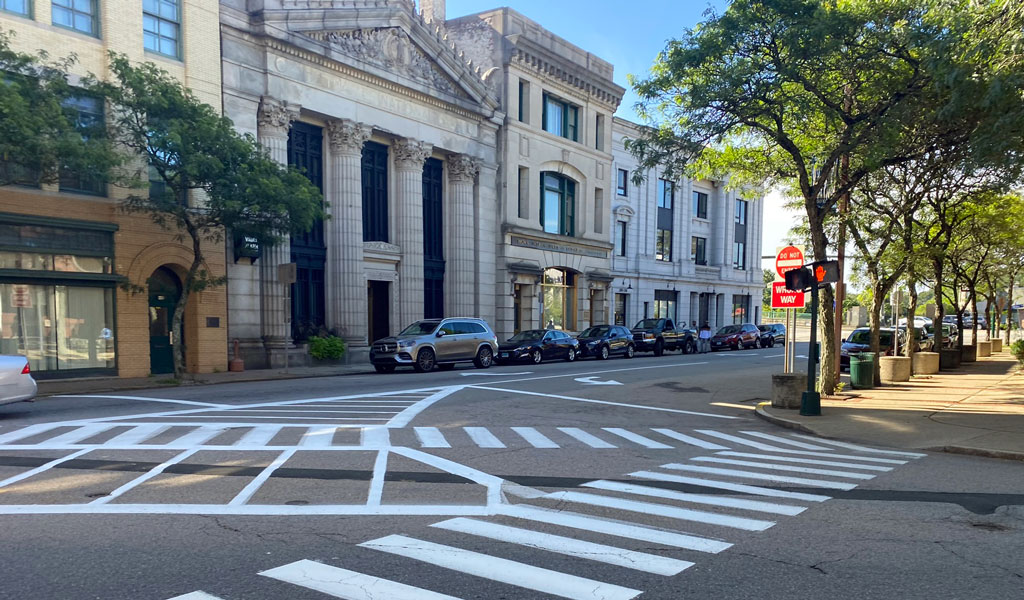 Street intersection with prominent white crosswalk markings, lined with trees, parked cars, and classic building architecture.  