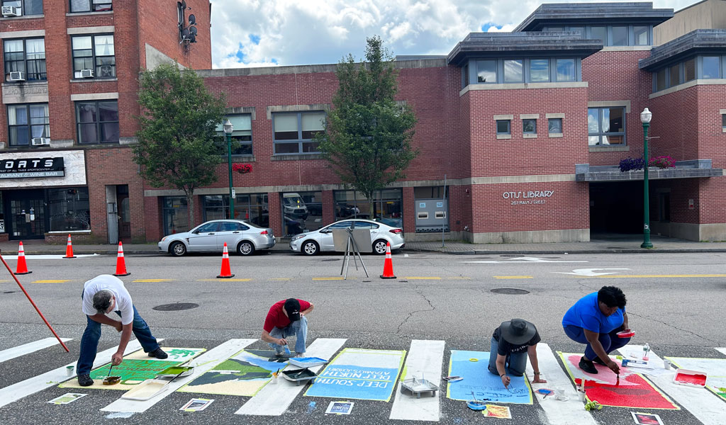A group of people paints a colorful street mural on a crosswalk across from a brick building labeled "O’Neill Library."