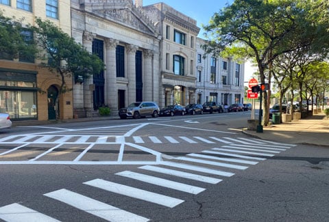 Street intersection with prominent white crosswalk markings, lined with trees, parked cars, and classic building architecture.  