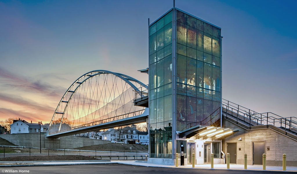 A glass elevator shaft, stairwell, and arched pedestrian bridge at sunset.