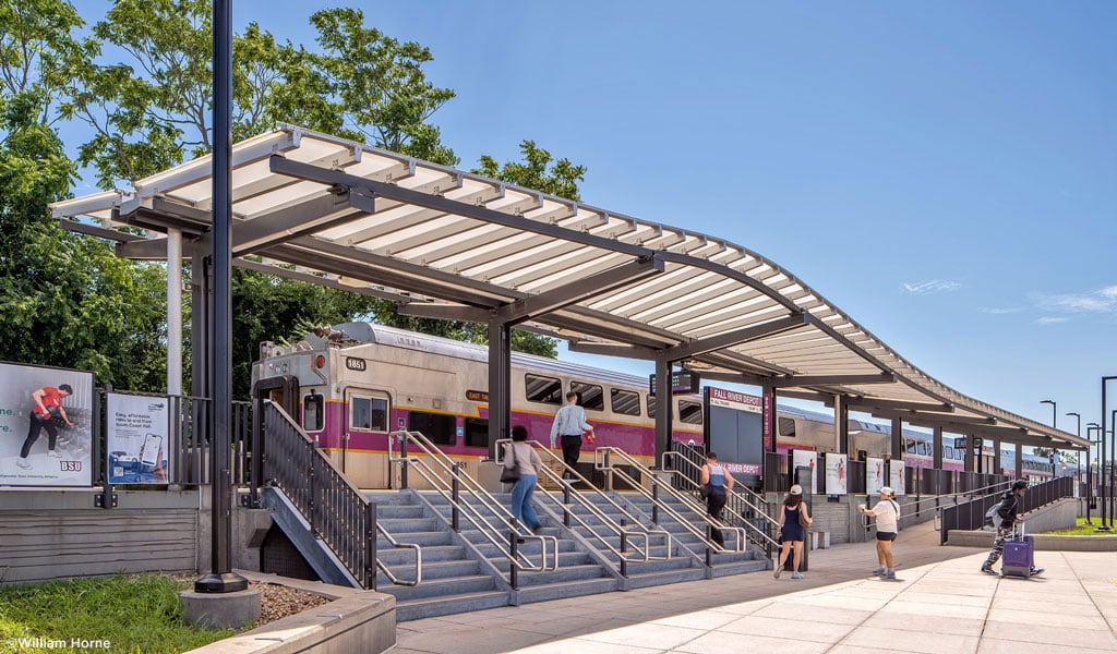 A train arriving at Fall River loading platform with covered station, stairwell, and wheelchair-accessible ramp.