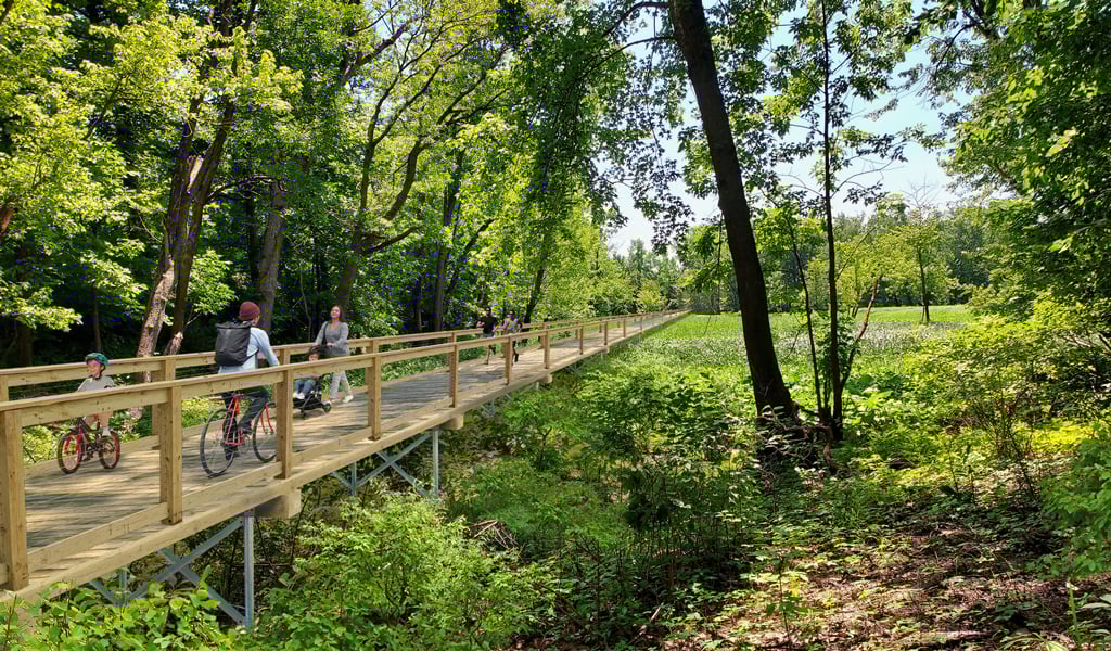 A proposed boardwalk running over a river showing bicyclists and pedestrians using the boardwalk through a wooded area.