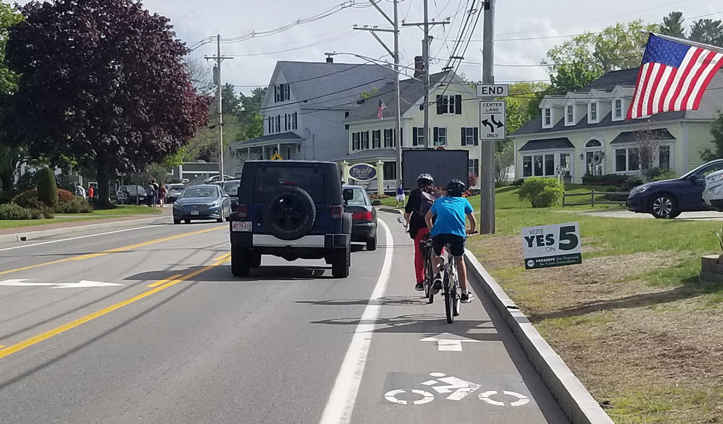 View of bike lane on Route 1 in Ogonquit, Maine, with two cyclists in the bike lane and multiple cars driving to their left.