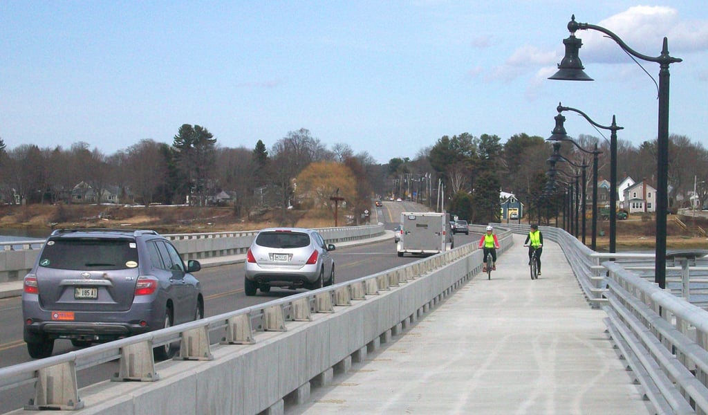 View of bike lane on Martin’s Point Bridge in Falmouth, Maine, with two cyclists in the bike lane and multiple cars driving to their left.