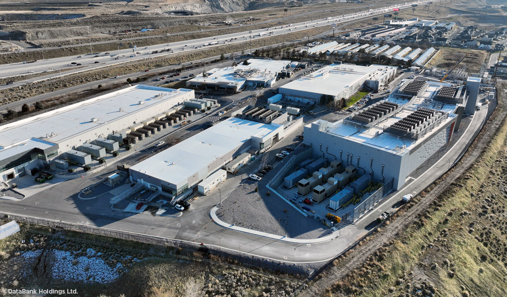 Aerial view of a large data center campus and backup generators alongside a highway and mountain range.
