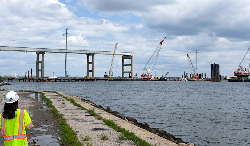 Woman in high-vis PPE observes Francis Scott Key Bridge site with cranes, pile supports and partial structure over water.