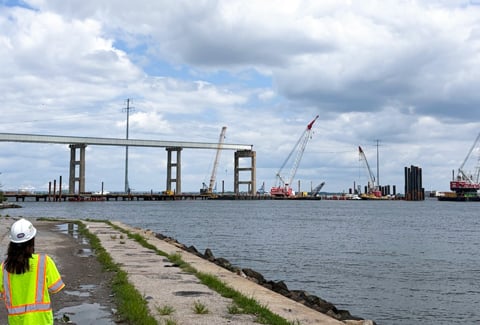 Woman in high-vis PPE observes Francis Scott Key Bridge site with cranes, pile supports and partial structure over water.