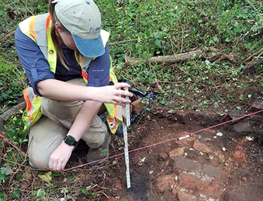 A woman kneels near a hole in the ground and measures the depth.