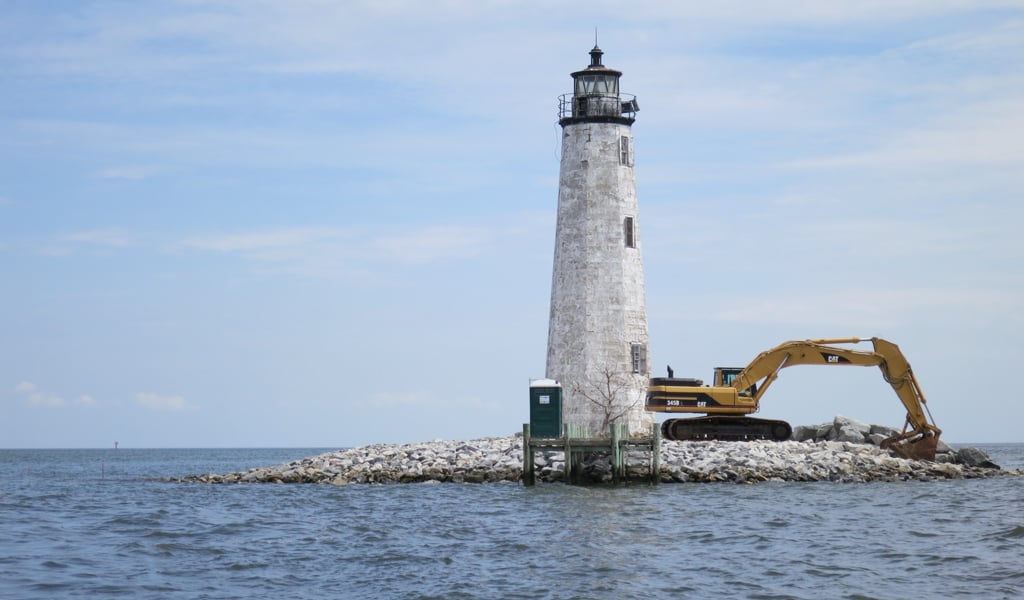 Lighthouse on a rocky island with an excavator beside it under a blue sky.
