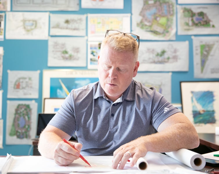  A man with blonde hair draws at a table with sketches hanging in the background.