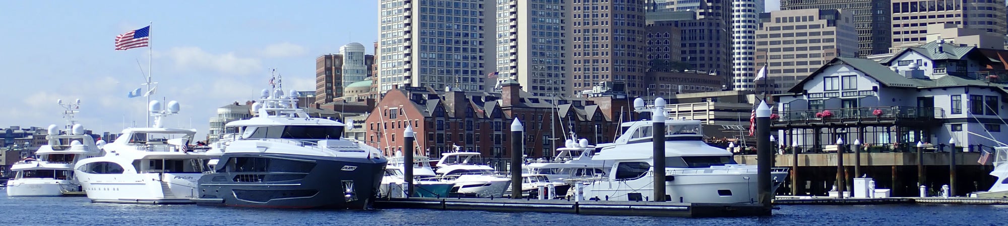 Luxury boats docked in a harbor with a city skyline in the background under a blue, partly cloudy sky.