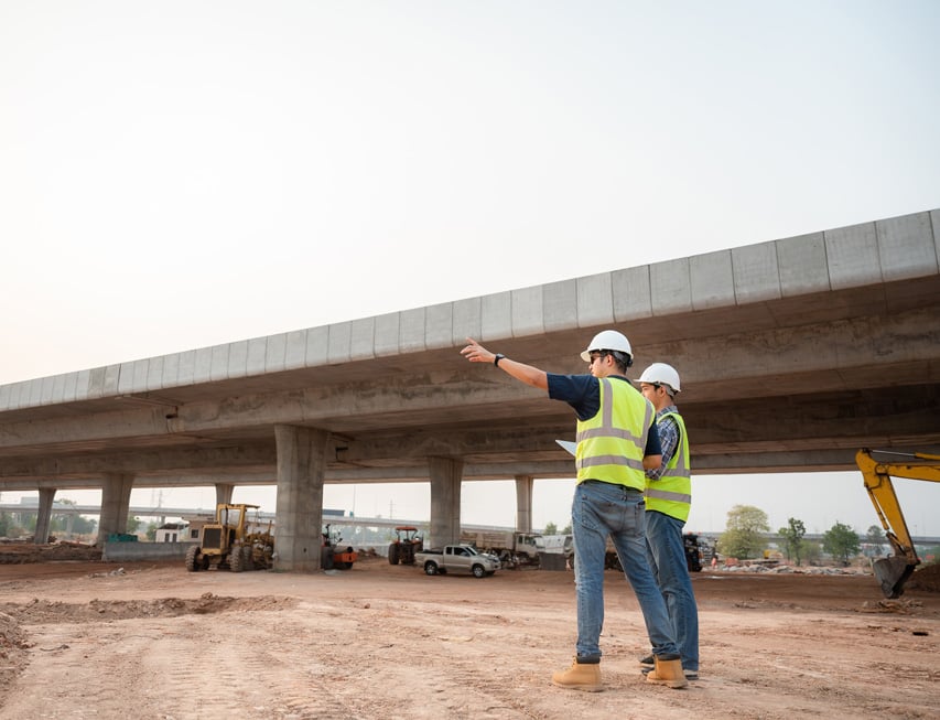 Workers in safety gear inspecting a highway construction site with vehicles and equipment in the background.