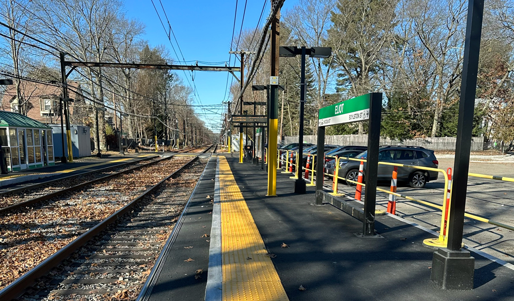 Outdoor train tracks with autumn leaves, a yellow tactile strip, overhead power lines, and a green station sign near parked cars.