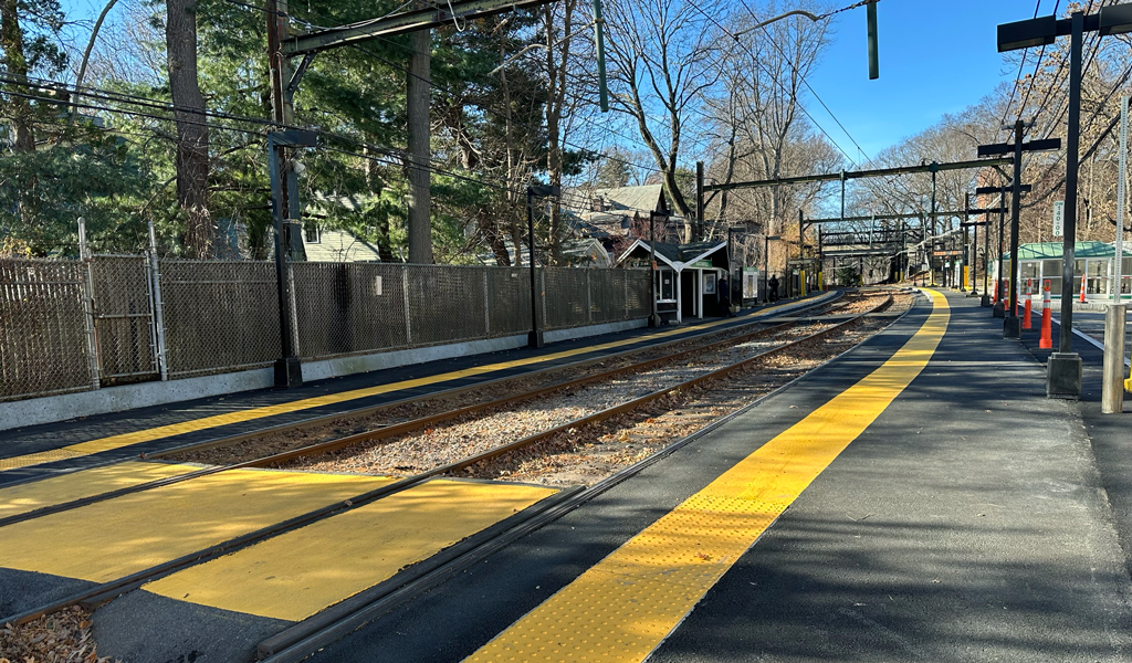 Train station platform with yellow tactile paving, bordered by tracks and overhead power lines, surrounded by trees.