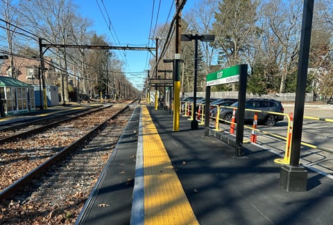 Outdoor train tracks with autumn leaves, a yellow tactile strip, overhead power lines, and a green station sign near parked cars.