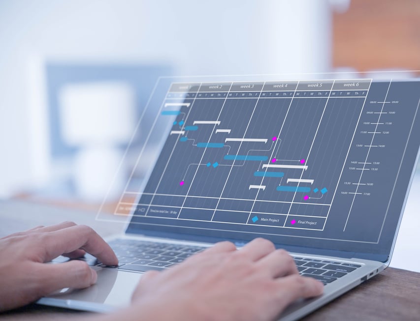 A person types on a laptop displaying a Gantt chart with project tasks and timelines in a modern, bright office setting.