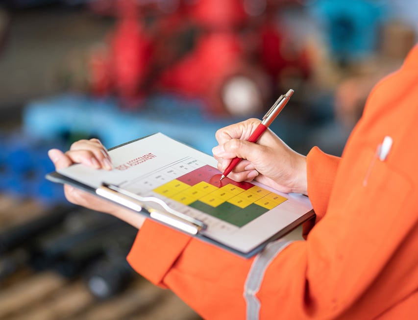 A person wearing an orange uniform writes on a risk assessment chart attached to a clipboard in an industrial setting.