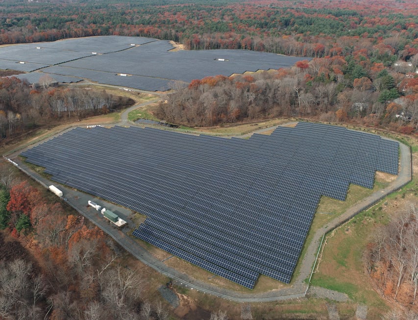 Aerial view of large solar farm surrounded by autumn-colored forested hills.