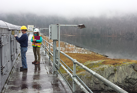Two people in hardhats stand on top of a dam and take measurements