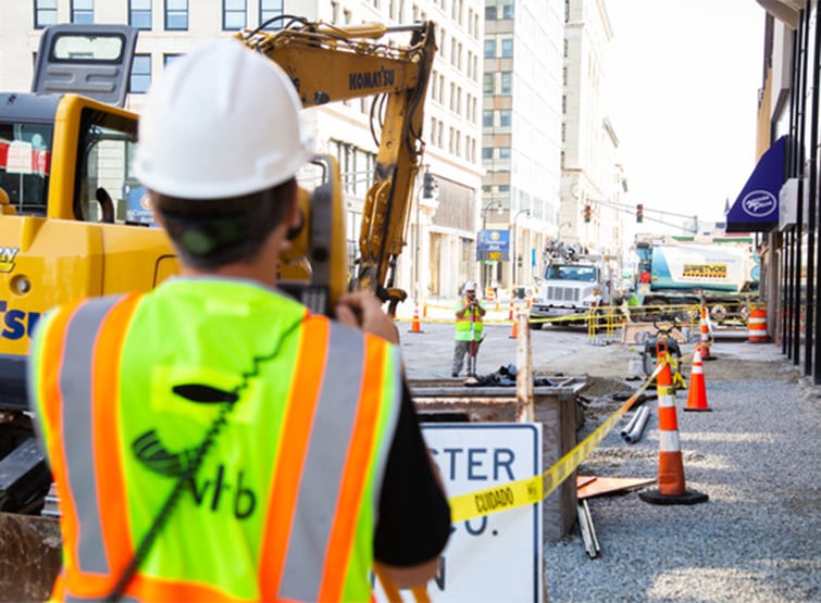 Man wearing white hard hat and bright safety vest operating survey equipment on city street. 