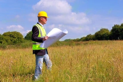 Man with construction hat and vest reading site plans in a field.