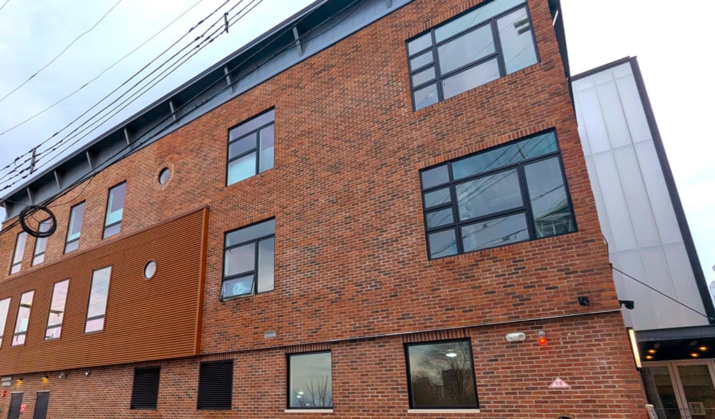 Brick waterfront residential building with large windows, landscaped foreground, and river visible beyond.
