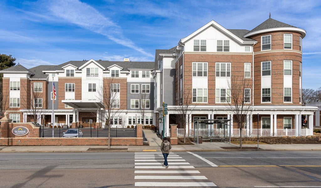 A four-story brick senior living facility under a clear blue sky with a pedestrian in the foreground crossing a four-lane road. 
