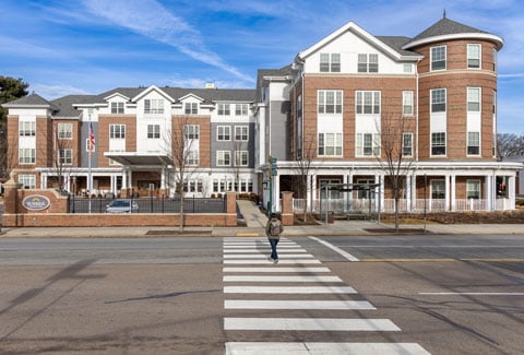 A four-story brick senior living facility under a clear blue sky with a pedestrian in the foreground crossing a four-lane road. 