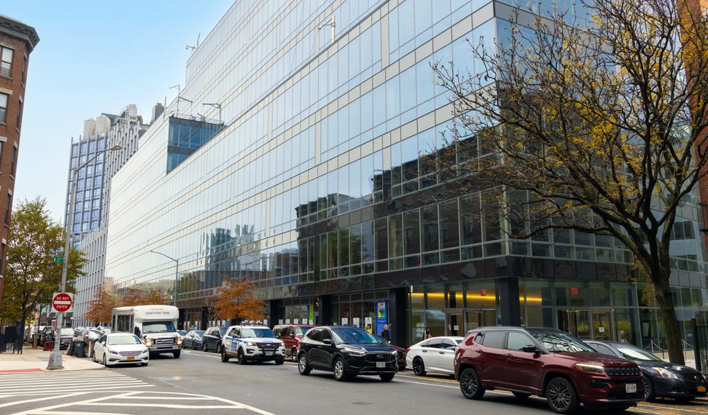 Modern glass lab building in New York with parked cars and traffic along a busy city street on a clear fall day.