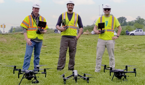 Three men stand in a grassy area behind three unmanned drones.