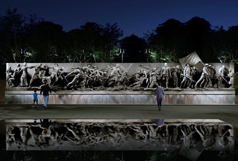 Long bronze wall sculpture of people in front of reflecting pool at night