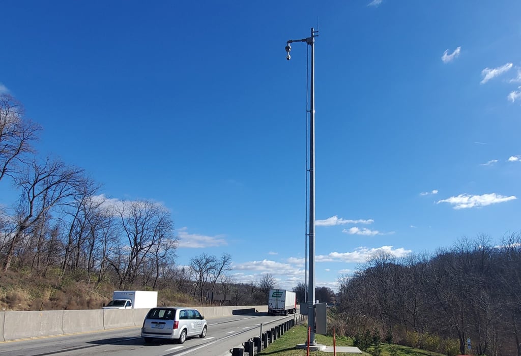 Traffic camera mounted on a tall pole along I-70 in Pennsylvania, with cars and trucks traveling on the highway.