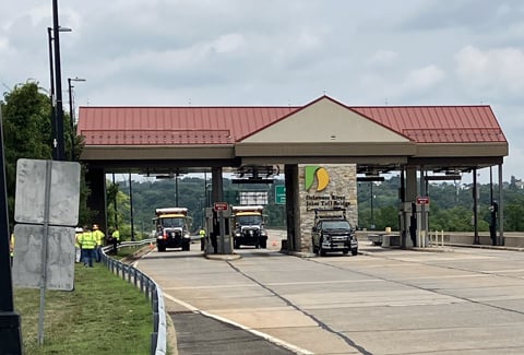 Trucks and construction workers near a toll plaza with red roof on a cloudy day.