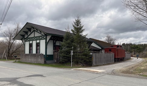 A pedestrian walks on a gravel trail alongside a historic train car and station.