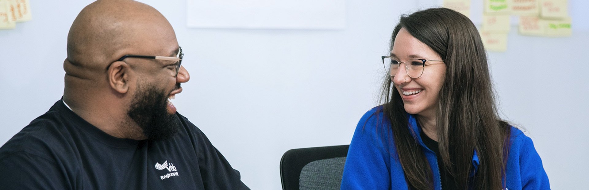 Two people sitting at a table with laptops, smiling at each other, with sticky notes on the wall behind them.