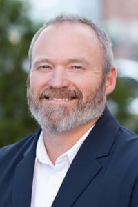 A person wearing a white button-down shirt and dark blazer is smiling outside with a tree in the background.