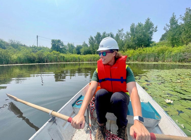 A person wearing a hard hat and life jacket is rowing a boat on a calm body of water surrounded by greenery.
