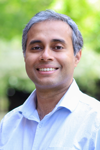 A person wearing a light button-down shirt is smiling outside with trees in the background.