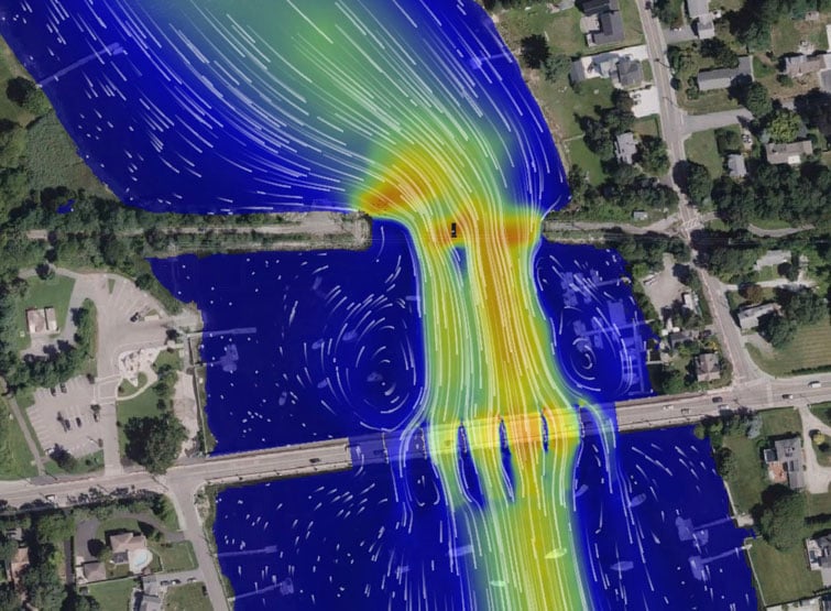 Aerial view of a suburban area featuring a river intersected by a bridge. A colorful overlay shows flow patterns over the water, with arrows indicating direction and intensity. 