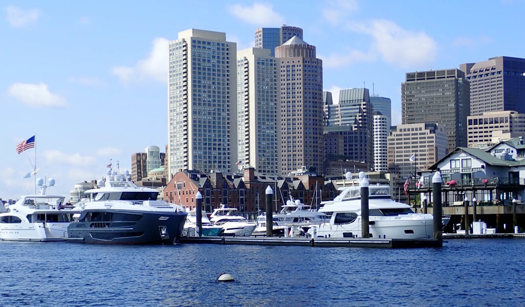 Luxury boats docked in a harbor with a city skyline rising behind them under a bright blue, partly cloudy sky.