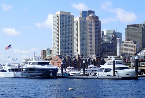 Luxury boats docked in a harbor with a city skyline rising behind them under a bright blue, partly cloudy sky.