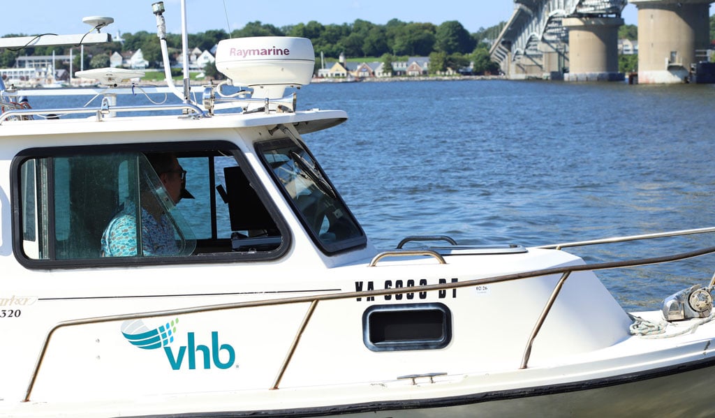 Person piloting a VHB boat on the water near a large bridge and shoreline homes.