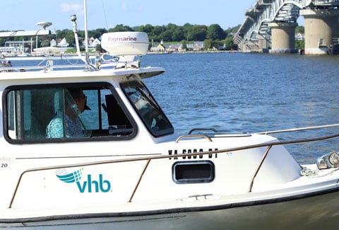Person piloting a VHB boat on the water near a large bridge and shoreline homes.